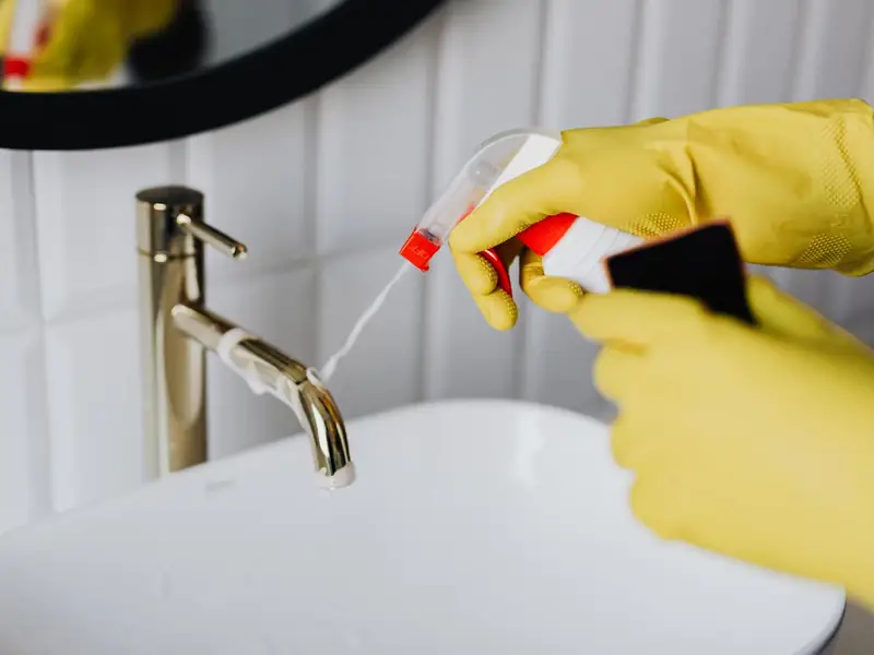 Gloved hands spraying disinfectant on a bathroom wash basin and tap